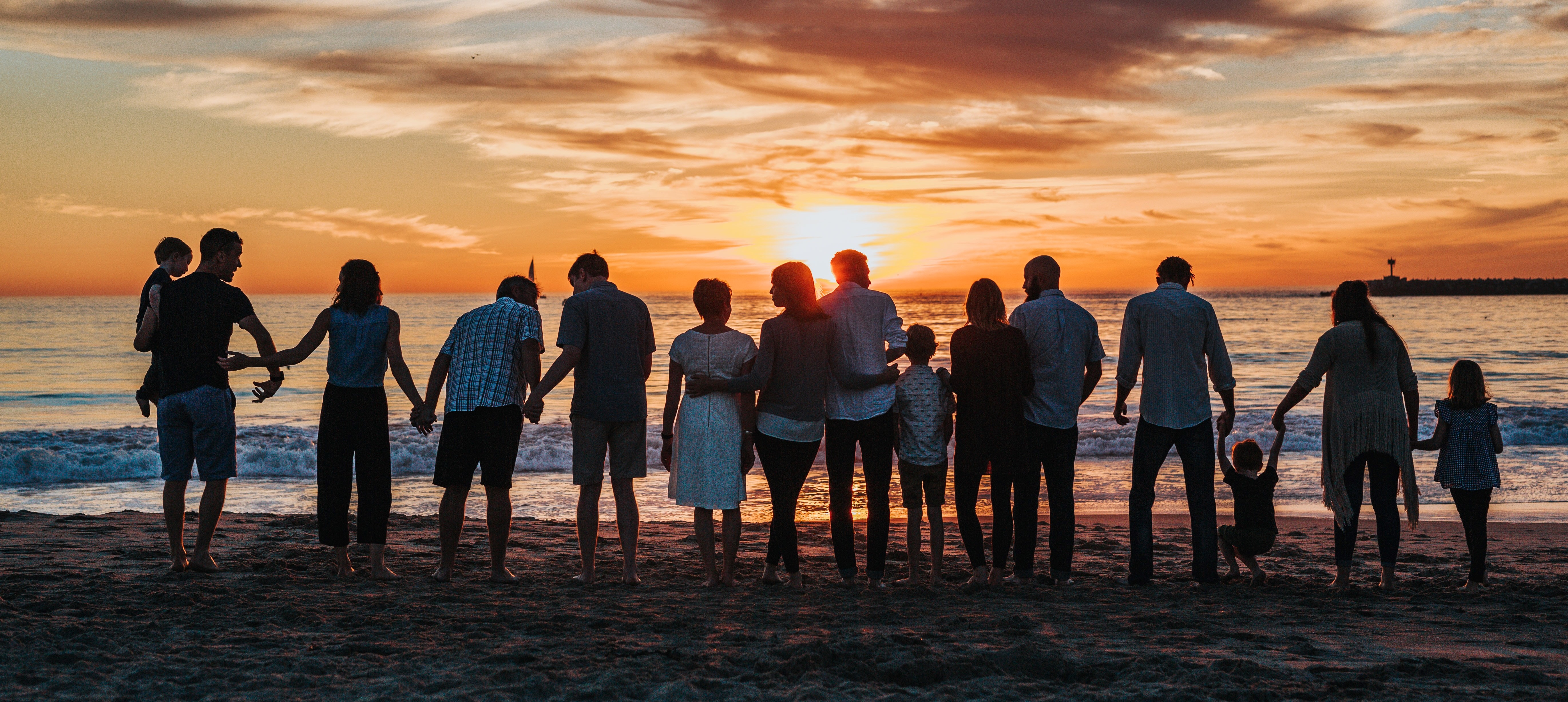 A group of adults of varying ages and genders standing on the beach, holding hands, and enjoying the beautiful sunset. In the distance, a stone breakwater with a small lighthouse can be seen. The image represents the importance of family bonds and creating a better business world with the Protopian philosophy.